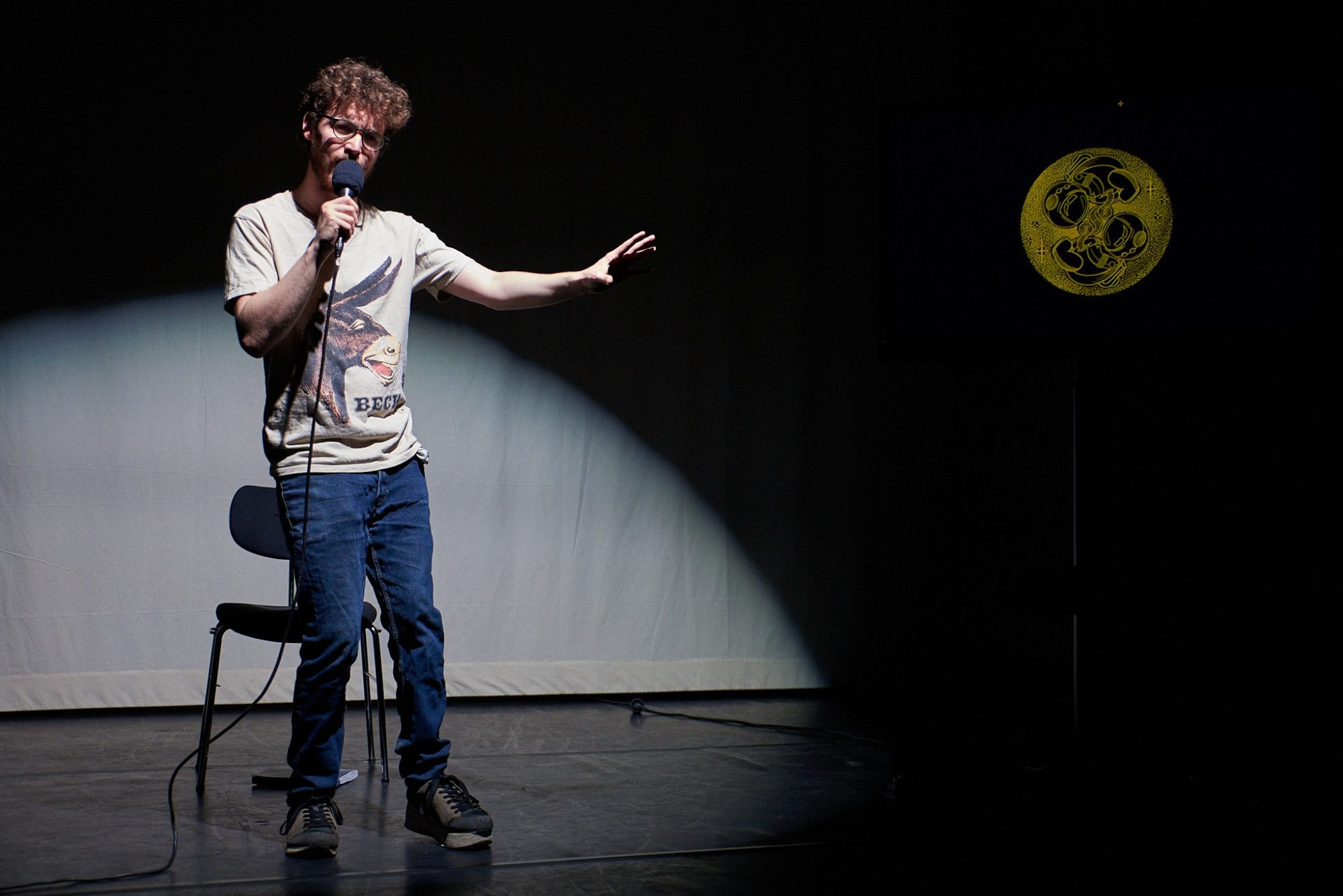 Jack stands in a spotlight, speaking into a microphone. He stands in front of a chair. The image of the two astronaut babies is shown in the darkness to the left of him.