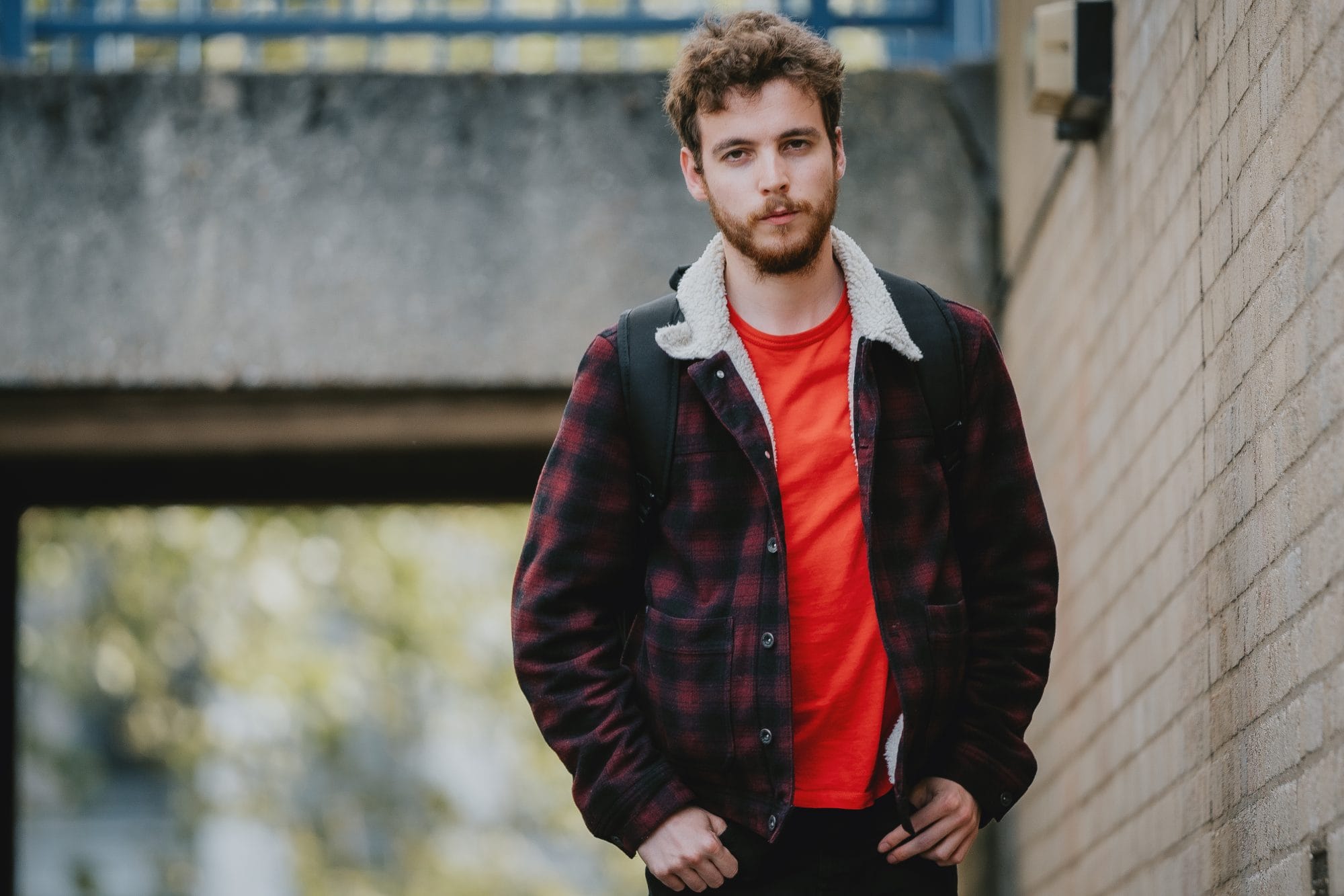 A man, wearing a red t and red and black checkered jacket, with a sheepskin collar walks past a concrete wall.