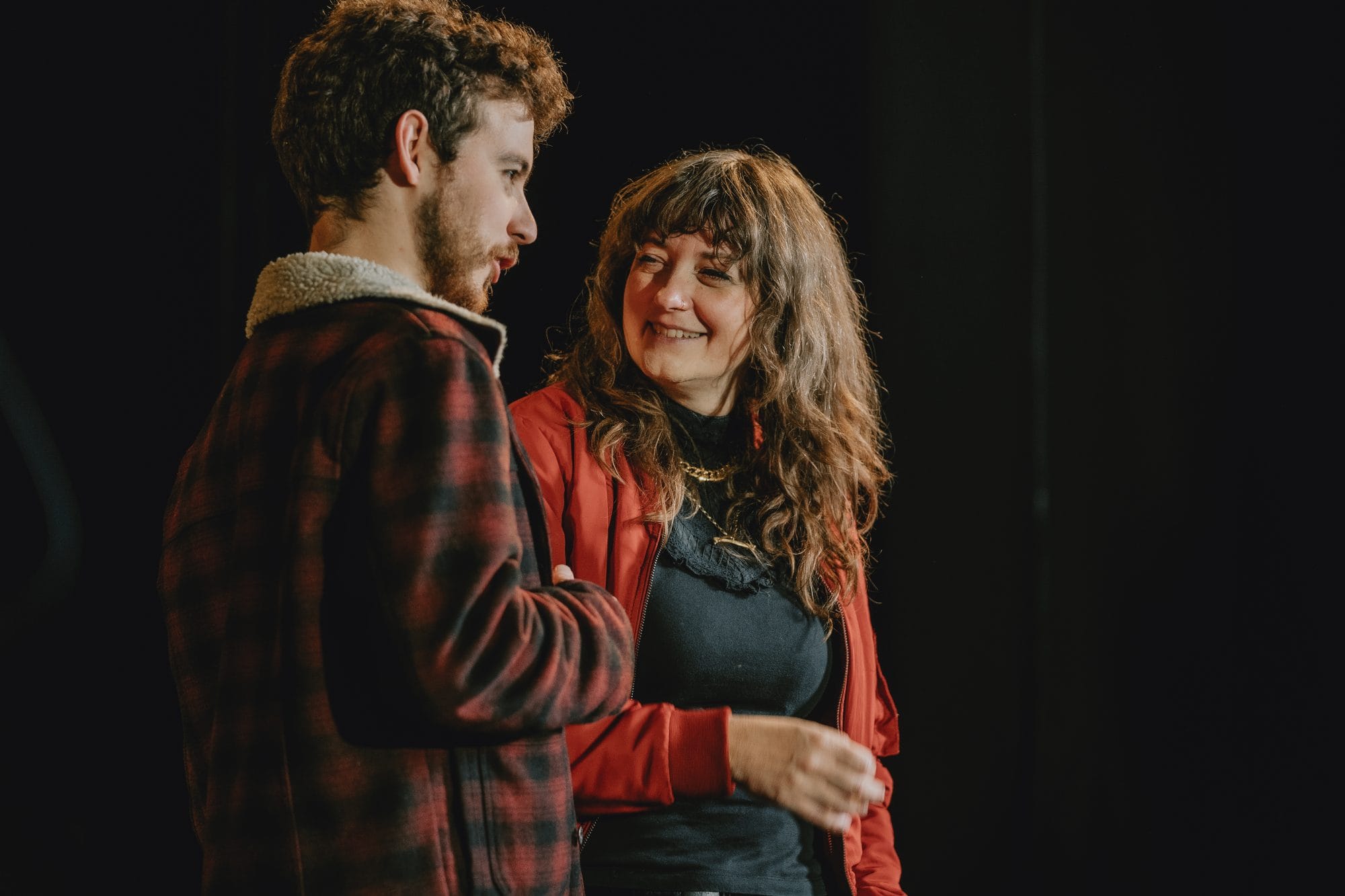 A man and woman, both wearing red and black are chatting and laughing. They are standing in a black box space.