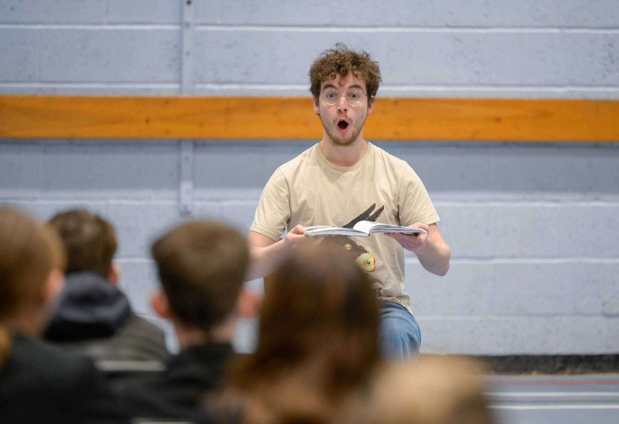 Jack sits on a chair in a school hall, with a surprised face. He is opening a book. School children watch him.