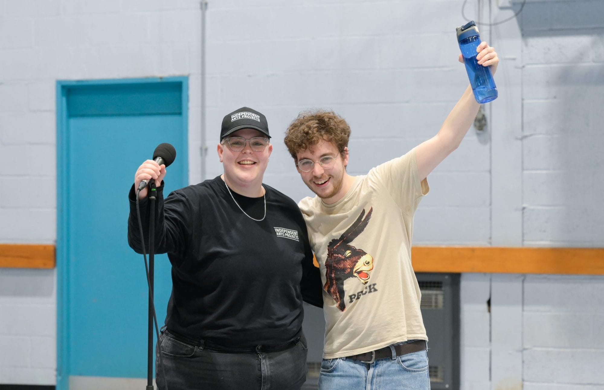 Jack stands with left arm out stretched in celebration, His right arm is around Susan. She is wearing a cap and holds onto the microphone stand.
