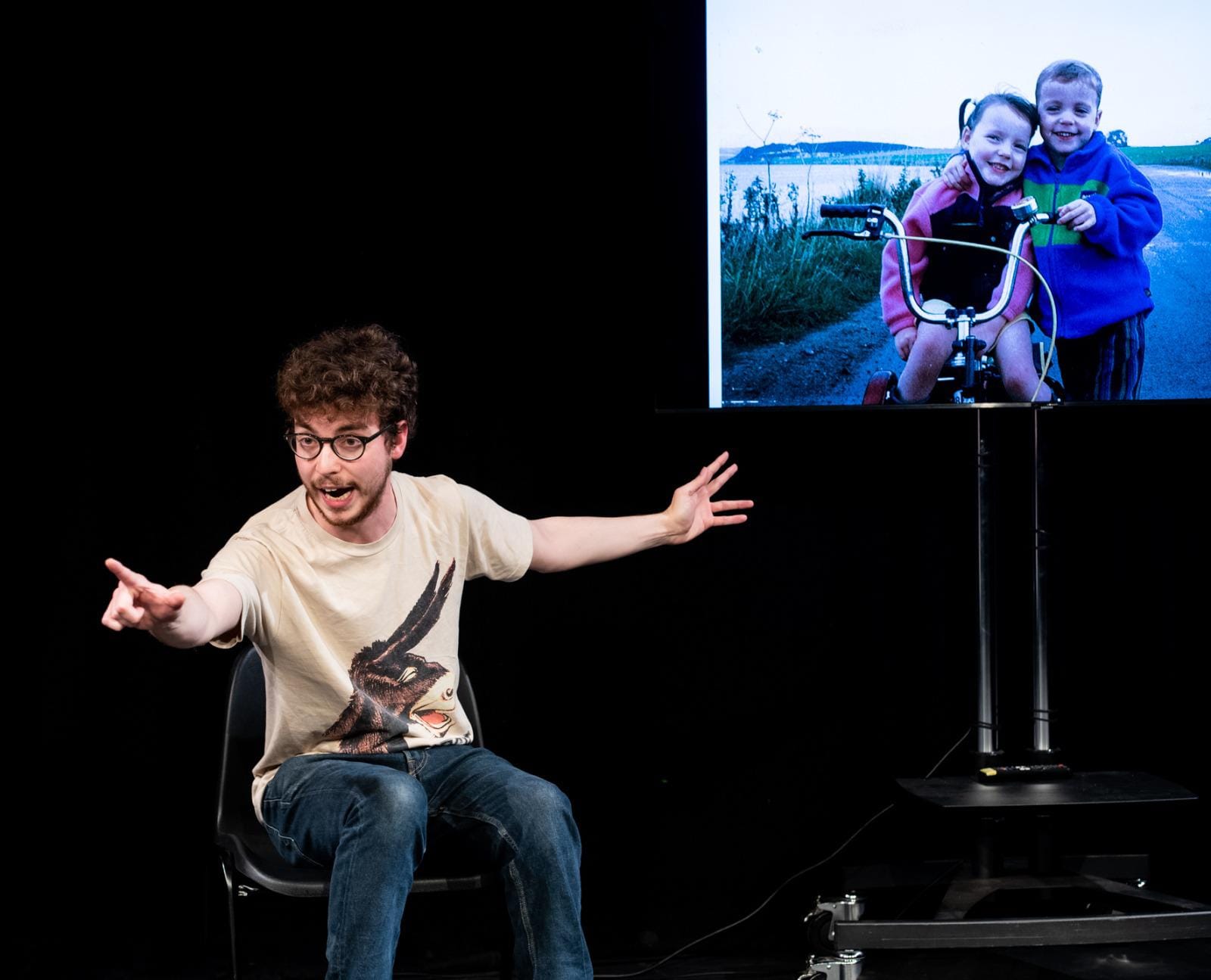 Jack sits on a chair and points at the audience. A screen, behind him, shows a picture of Bec and him as children.