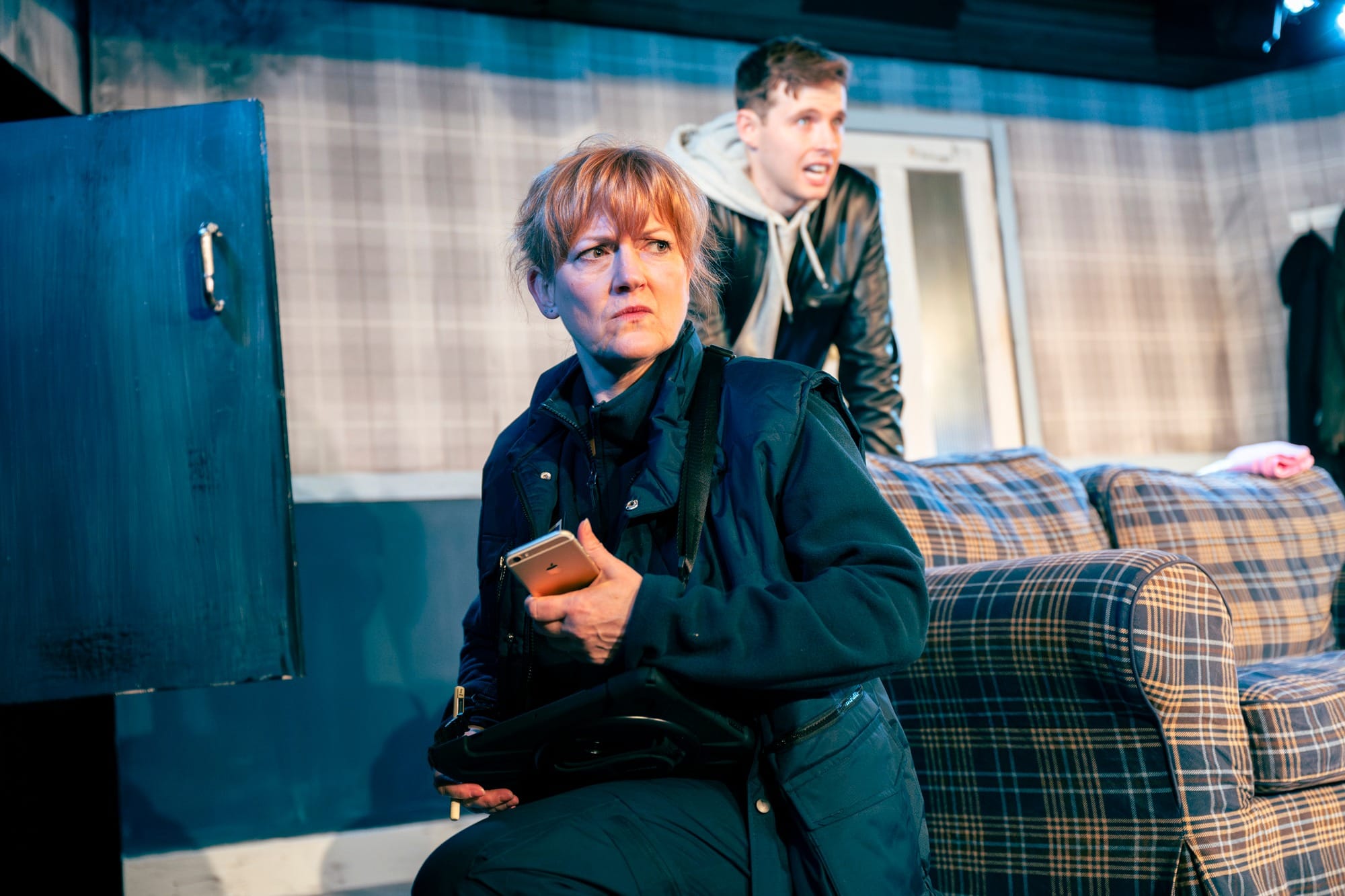 A red headed woman, kneels beside a sofa. She is holding a phone. A man leans on the back of the sofa, behind her.