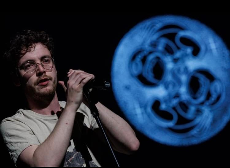 Jack stands at a mic stand, with both hands on the microphone. The image of the two astronaut babies looms behind him, like the moon.
