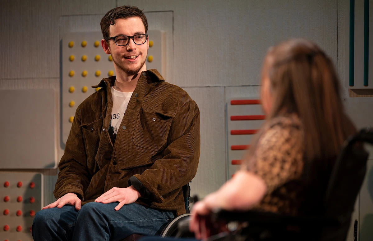 A young man, smiling in a wheelchair. Curly hair, glasses. He is wearing a brown Jacket and blue jeans. A young woman in a wheelchair is facing him. She is wearing a leopard print top and has long brown hair.