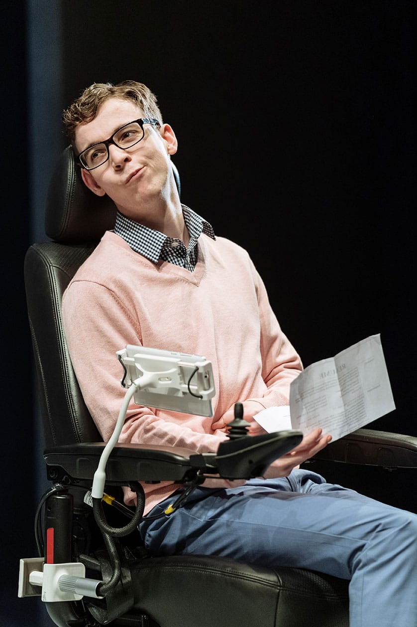 A man in a wheelchair holds a letter and looks on with a smug face. He is wearing s pink jumper, with a blue shirt collar and trousers. A tablet stand is locked to the arm rest.