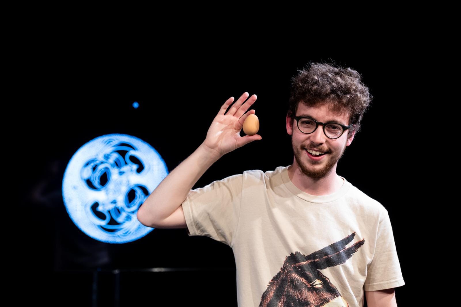 Jack holds an egg in his right arm, up to his face. Behind is the image of two astronaut babies in the womb.