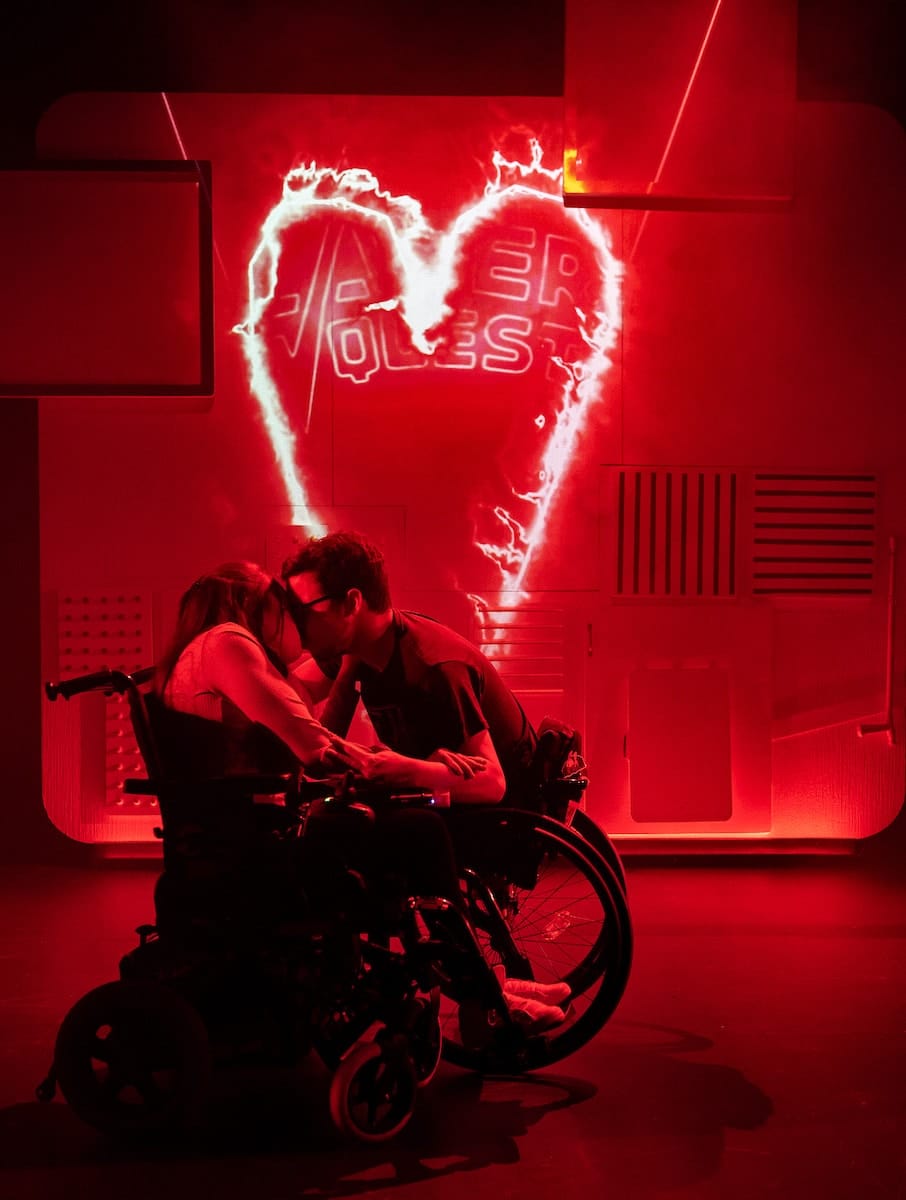 A man and woman in wheelchairs kiss passionately. The lighting is red. Behind them projected on the wall is a red lighting love heart. Inside the love heart. Text reads: Laser quest.
